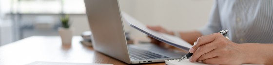 A person sitting at their laptop takes notes with a pen and paper
